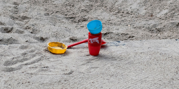 In einem Sandkasten liegen eine gelbe Schaufel, ein rotes Eimerchen und ein blauer Sandtrichter