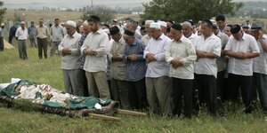 Männer beten vor einer eingehüllten Leiche, die auf einer Bahre liegt.