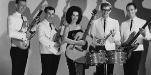 Die junge Frontfrau Wanda Jackson steht mit ihrer Gitarre in der Mitte. Rechts und links von ihr stehen jeweils zwei Männer mit ihren Instrumenten. Alle lächeln.