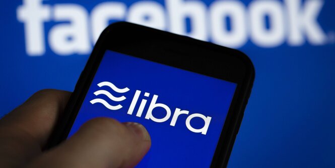 Jemand hält ein Handy, auf dem das Libra-Logo angezeigt wird. Im Hintergrund ist das Logo von Facebook zu sehen