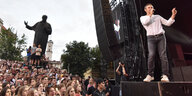 Ein Mann in weißem Shirt steht auf einer Bühne, vor ihm viele Menschen, im Hintergrund eine Statue in ähnlicher Haltung