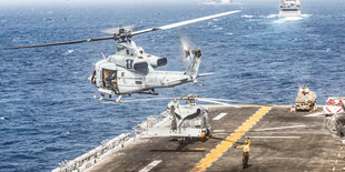 Ein Erkundungshubschrauber hebt vom Deck des Kriegsschiff USS Boxer ab. Das Bild ist aus der Vogelperspektive aufgenommen. Ein Lotse steht unten und winkt dem Hubschrauber zu.