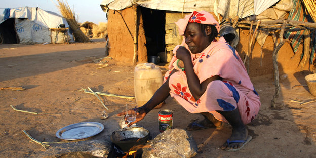 Eine Frau sitzt vor einem Lehmhütte und bereitet auf einer kleinen Kochstelle etwas zu essen vor.
