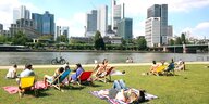 Leute liegen auf dem Rasen oder sitzen in einem Liegestuhl am Ufer des Mains - gegenüber die Hochhauskulisse von Frankfurt