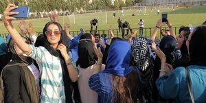 Frauen mit Kopftüchern schauen durch einen Gitterzaun, dahinter sieht man ein Fußballspiel, eine Frau macht ein Selfie von sich