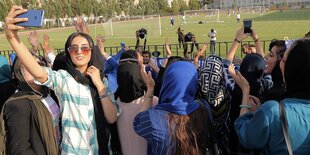 Frauen mit Kopftüchern schauen durch einen Gitterzaun, dahinter sieht man ein Fußballspiel, eine Frau macht ein Selfie von sich