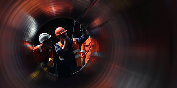 Drei Arbeiter mit orangenem Schutzanzug und Schutzhelm schauen in eine Röhre einer Gas-Pipeline rein.