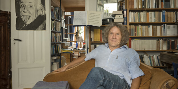 Günther Egidi sitzt auf einer Couch im Hintergrund ein Bücherregal und eine offene Tür, die in ein Arbeitszimmer führt. An der Tür hängt das berühmte Plakat von Albert Einstein mit herausgestreckter Zunge
