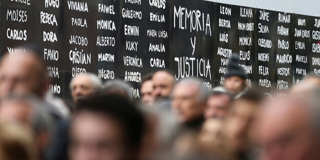 Im Hintergrund sieht man eine schwarze Tafel, auf der in weißer Schrift viele Vornamen zu lesen sind. Davor verschwommen eine Menschenmenge.