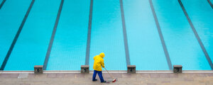 Mitarbeiter mit gelber Jacke fegt neben leerem Schwimmbecken