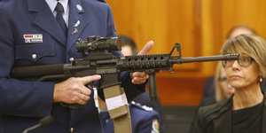 Im Gerichtssaal: Ein Mann in Polizeiuniform hält ein Gewehr und zeigt es einer Frau.