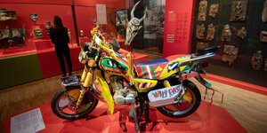 Ein Motorrad steht auf einem Podest im Museum. Es ist bunt bemalt und hat Fransen am Lenker