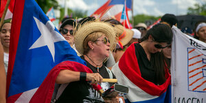Auf dem Bild eines Protests in Puerto Rico ist links die Flagge des Territoriums zu sehen, in der Mitte eine Frau mit Strohhut, die mit einem Stick auf eine Kuhglocke schlägt.
