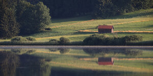 Eine Hütte spiegelt sich in einem See, sie steht einsam auf einer weiten Wiese