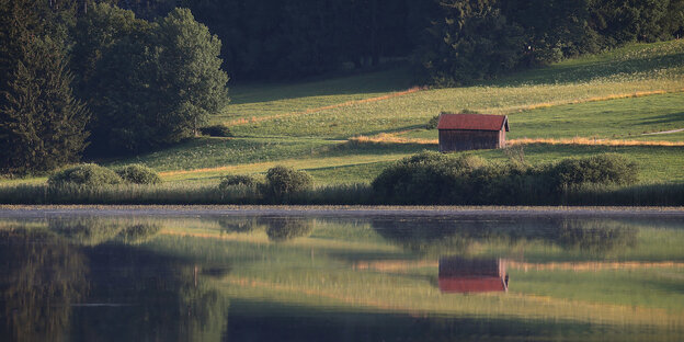 Eine Hütte spiegelt sich in einem See, sie steht einsam auf einer weiten Wiese