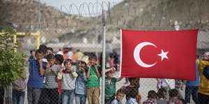 Geflüchtete Kinder warten in einem Lager hinter einem Stacheldrahtzaun, auf dem die türkische Flagge prangt.