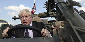 Boris Johnson steuert ein Militärfahrzeug. Er hält das Lenkrad fest, hat Augen und Mund weit aufgerissen. Im Hintergrund wehen die britische und die polnische Fahne.