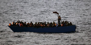 Flüchtende Menschen auf einem kleinen Boot im Mittelmeer. Einer steht und wedelt mit einem T-Shirt, um nach Hilfe zu rufen.