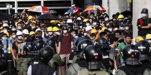 Eine Gruppe von Demonstranten steht in Hongkong Polizisten gegenüber. Die Demonstranten tragen Bauarbeiter- und Fahrradhelme sowie Mundschutz. Einige haben Regenschirme dabei.