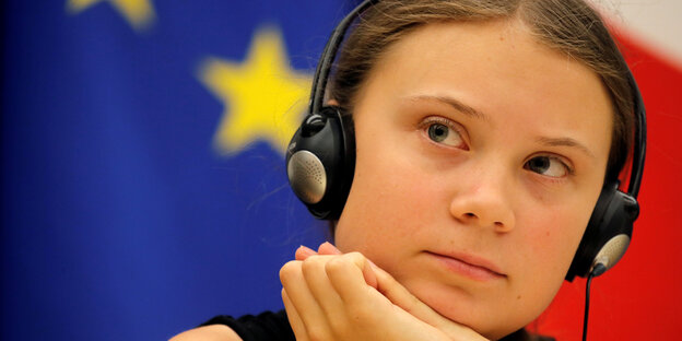 Greta Thunberg vor der EU-Flagge mit Kopfhörern in nachdenkender Pose