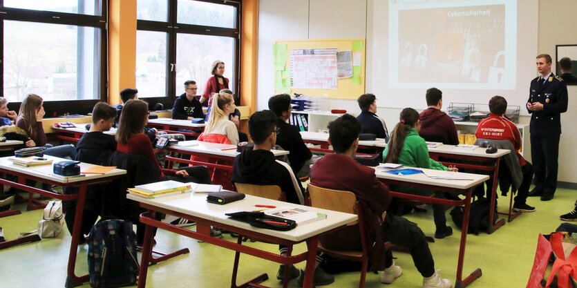 Ein Klassenraum mit vielen Tischen, Stühlen und Schüler*innen. Vorne rechts steht ein Jugendoffizier