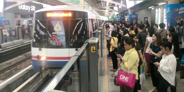 Zug in Bangkok fährt an Bahnsteig ein. Wartende Passagiere werden durch Glaswände vom Gleisbett getrennt