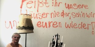 eine alte Frau sitzt in einem karg möblierten Zimmer auf einem Sofa, an die Wand wurde mit roter Farbe gesprüht: „Reißt ihr unsere Häuser nieder, seh'n wir uns in euren wieder!“