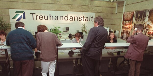 Menschen stehen mit Formularen an einem Bürothresen, hinter dem Sachbearbeiterinnen sitzen. Im Hintergrund an der Wand steht das Wort "Treuhandanstalt".