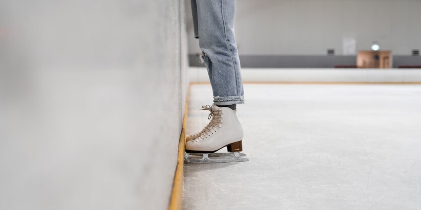 Blick auf die Schlittschuhe von jemandem, der an die Wand in einer EIslaufhalle.