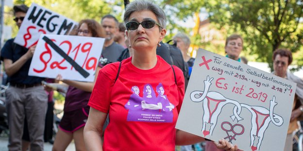 Eine Frau im roten T-shirt hält sichtbar genervt ein Schild "es ist 2019" in die Höhe
