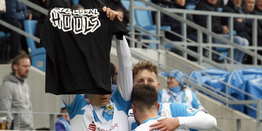 Daniel Frahn, in Fußballtrikot auf dem Feld neben anderen Spielern, hält ein T-shirt mit der Aufschrift "support your local hools" hoch