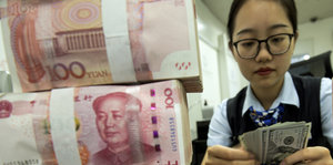 Links große chinesische rötliche Geldscheine gestapelt, links eine Bankmitarbeiterin, die weitere Scheine zählt