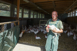 Ein Mann in grüner Latzhose trägt sein weißes Huhn durch den Stall