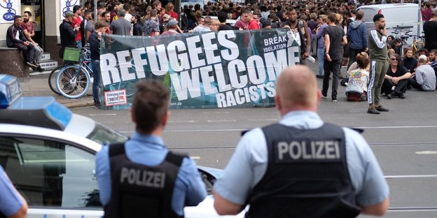 Polizisten stehen vor einer Demonstration, die Demonstranten halten einen Banner, auf dem steht: Refugees Welcome
