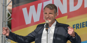 Björn Höcke vor einem Plakat in Deutschlandfarben, auf dem "Wende 2.0" steht. Er hält eine Rede und hebt dabei dir Arme.