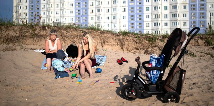 Zwei Frauen und ein Kind sitzen an einem Sandstrand, im Hintergrund ist ein Plattenbauriegel zu sehen, neben ihnen: ein Kinderwagen