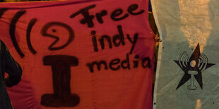 Transparente bei einer Demonstration. Auf dem einen steht "Free indymedia". Es trägt das Zeichen von linksunten.indymedia, ein i, das von Klammern umgeben ist. Auf dem anderen Transparent ist nur das Zeichen und darunter ein Stern.