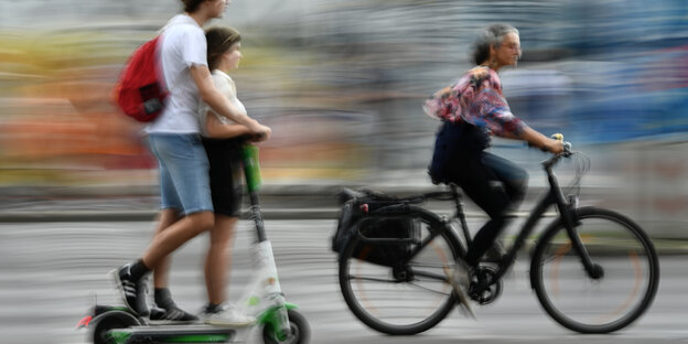 Eine Radfahrerin fährt an zwei Personen auf einem E-Scooter vorbei