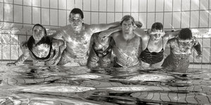 Schwarz-Weiß-Fotografie zeigt mehrere Menschen, in Badekleidung, die sich in einem Schwimmbecken stehend umarmen und die Augen zusammenkneifen