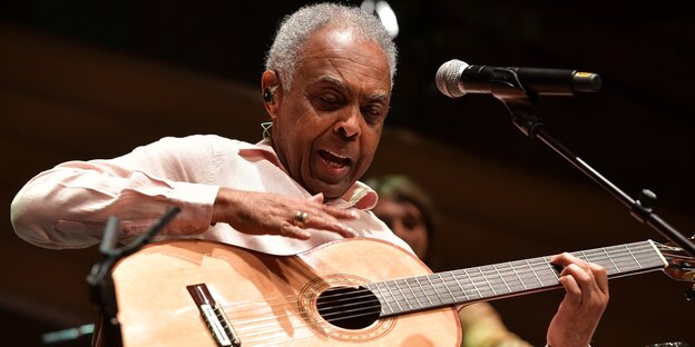 Gilberto Gil sitzt vor einem Mokrofon. Der alte Mann mit den kurzen Haaren singt und spielt dabei Gitarre.