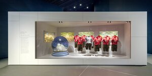 Im Museum für Bayerische Geschichte steht eine große Schneeglaskugel mit Schloss Neuschwanstein darin und daneben Lederhossen der Spieler vom 1.FC Bayern. Im HIntergrund sind Bilder vom Oktoberfest in München