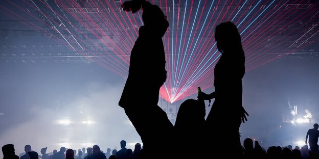 Silhouetten zweier tanzender Menschen, im Hintergrund Scheinwerfer und andere Tanzende