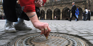 Eine Hand wirft eine Münze in einen Gulli vor dem Bremer Rathaus, der als Spendenbox dient