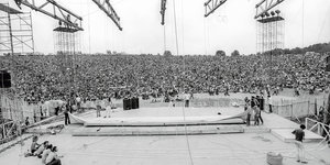 Im Vordergrund: Arbeiter beim Bühnenaufbau in Woodstock. Im Hintergrund: Eine Menschenmenge auf einem Feld