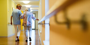 Eine Pflegekraft geht in einem Pflegeheim mit einer älteren Dame über einen Korridor.