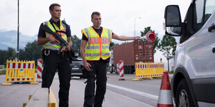 Zwei Polizisten halten einen weißen Wagen an. Sie tragen Warnwesten, einer hält eine Schusswaffe