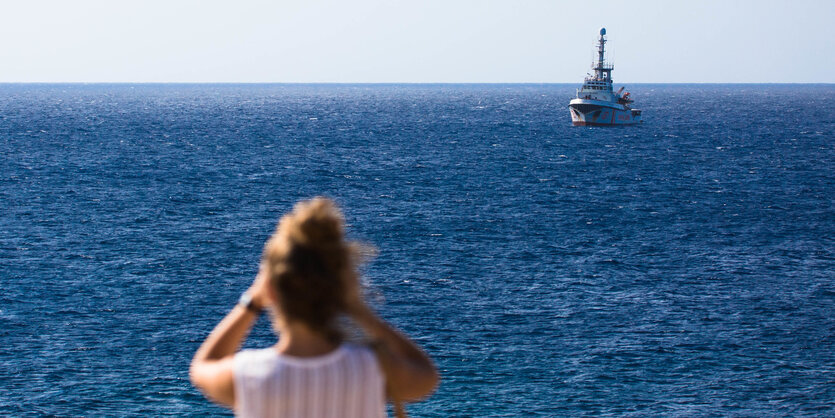 Eine Frau an Land guckt mit einem Fernrohr nach dem SChiff "Open Arms", das vor der Küste liegt