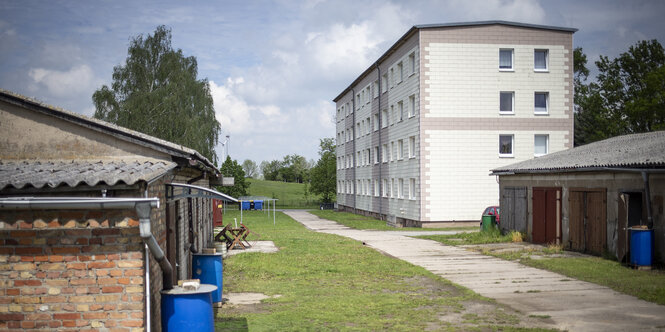 Ein weißer Flachbau steht neben alten Schuppen, dazwischen eine grüne Wiese