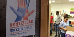 Im Vordergrund ein Unteilbar-Demo-Plakat. Im Hintergrund: Geflüchtete, die Plakate basteln