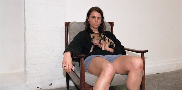 Die Musikerin Hackmann sitzt auf einem Sessel, blickt selbstbewusst in die Kamera und hält ein Bebi Schwein auf dem Arm
